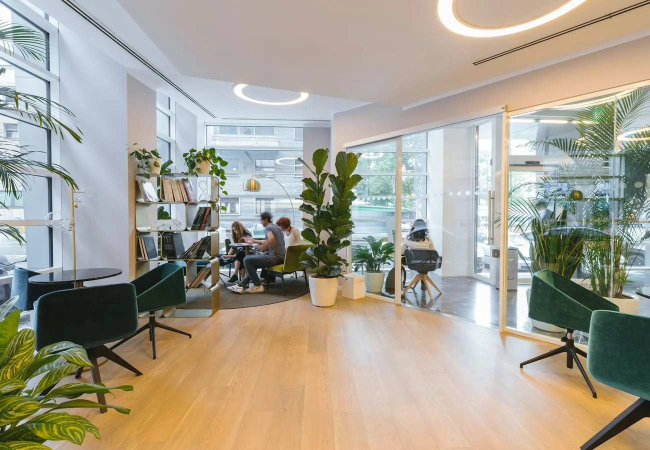 A bright workspace lounge with plants, armchairs and people working at the far tables