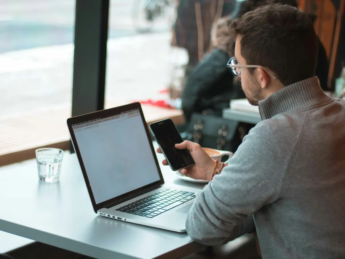 A man working on a laptop with his phone in hand at a window table