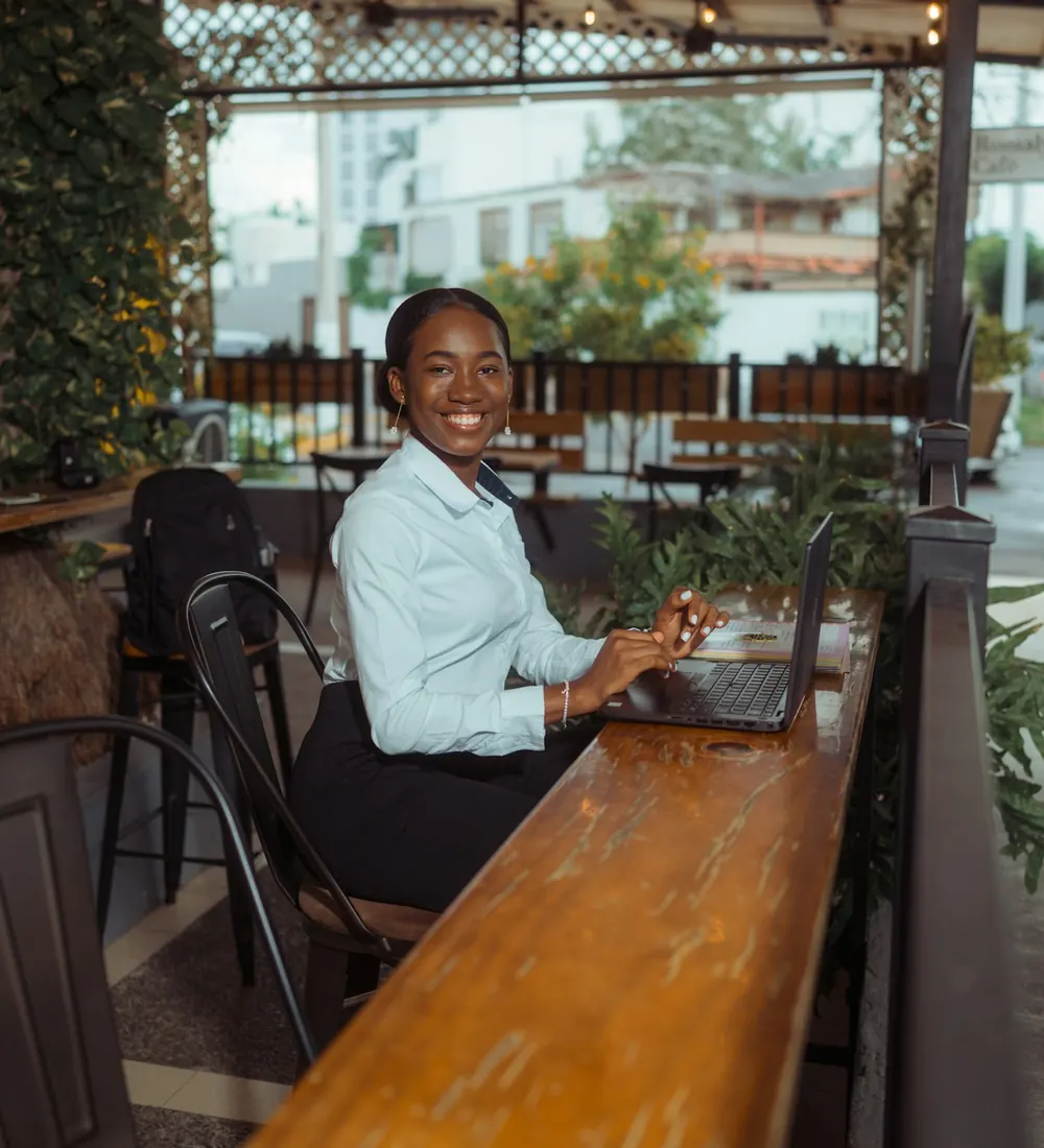 A smiling woman working on a laptop at a long counter on a covered terrace